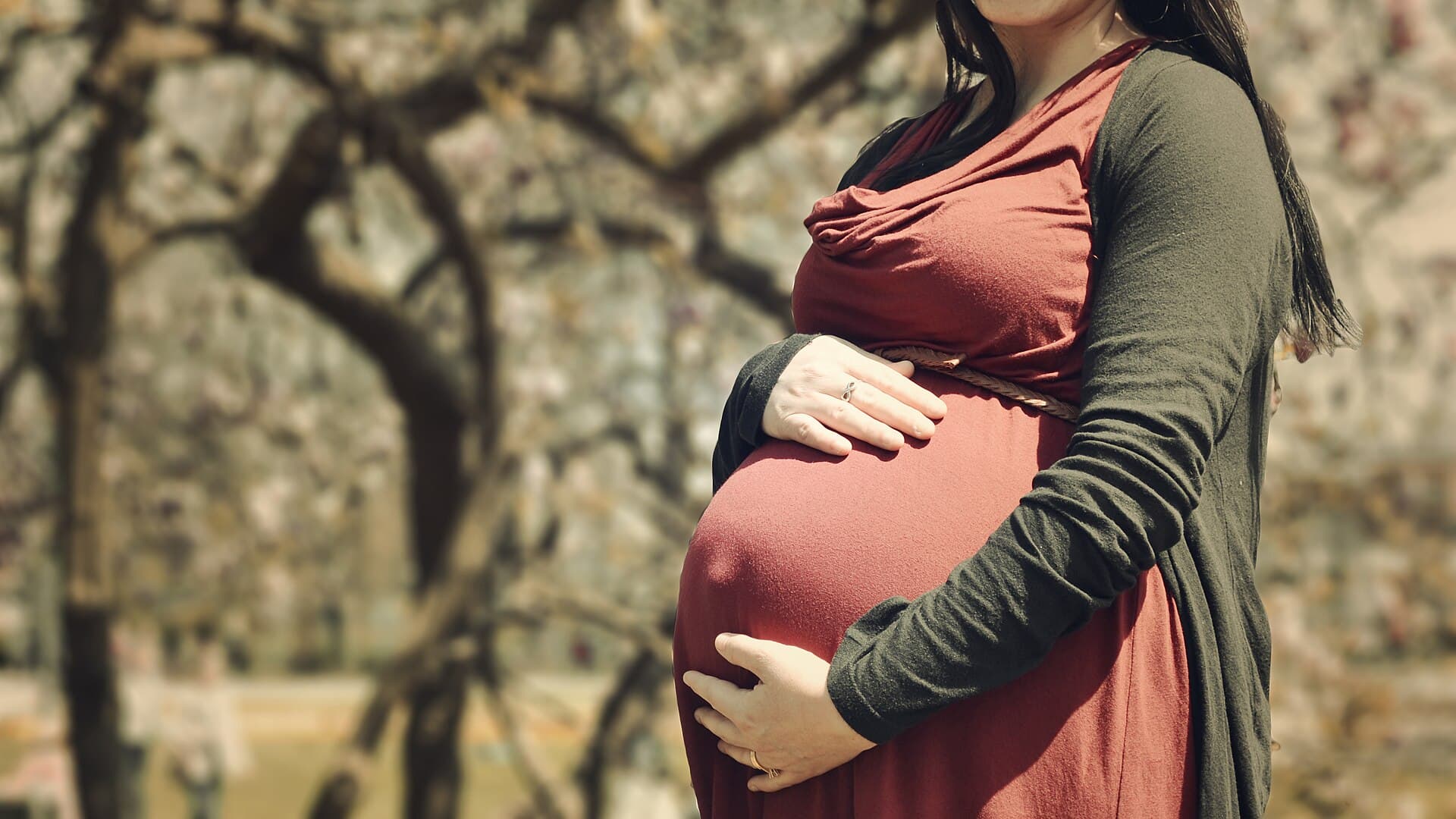 Zwangere vrouw in een rode jurk met een groen vest, met haar handen op haar buik, tussen herfstbomen