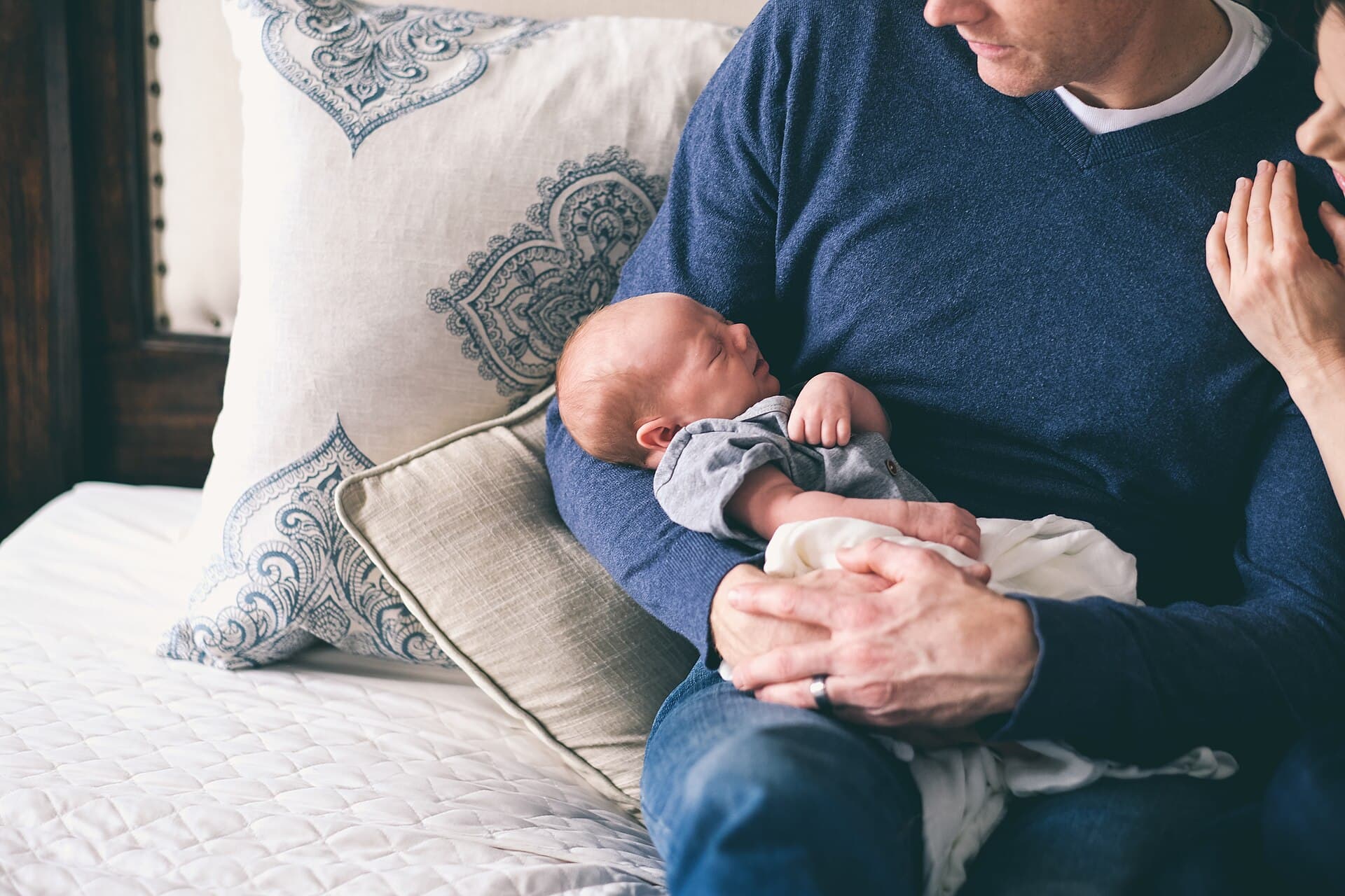 Vader zit op een bed met een pasgeboren baby in zijn armen, in een lichte slaapkamer