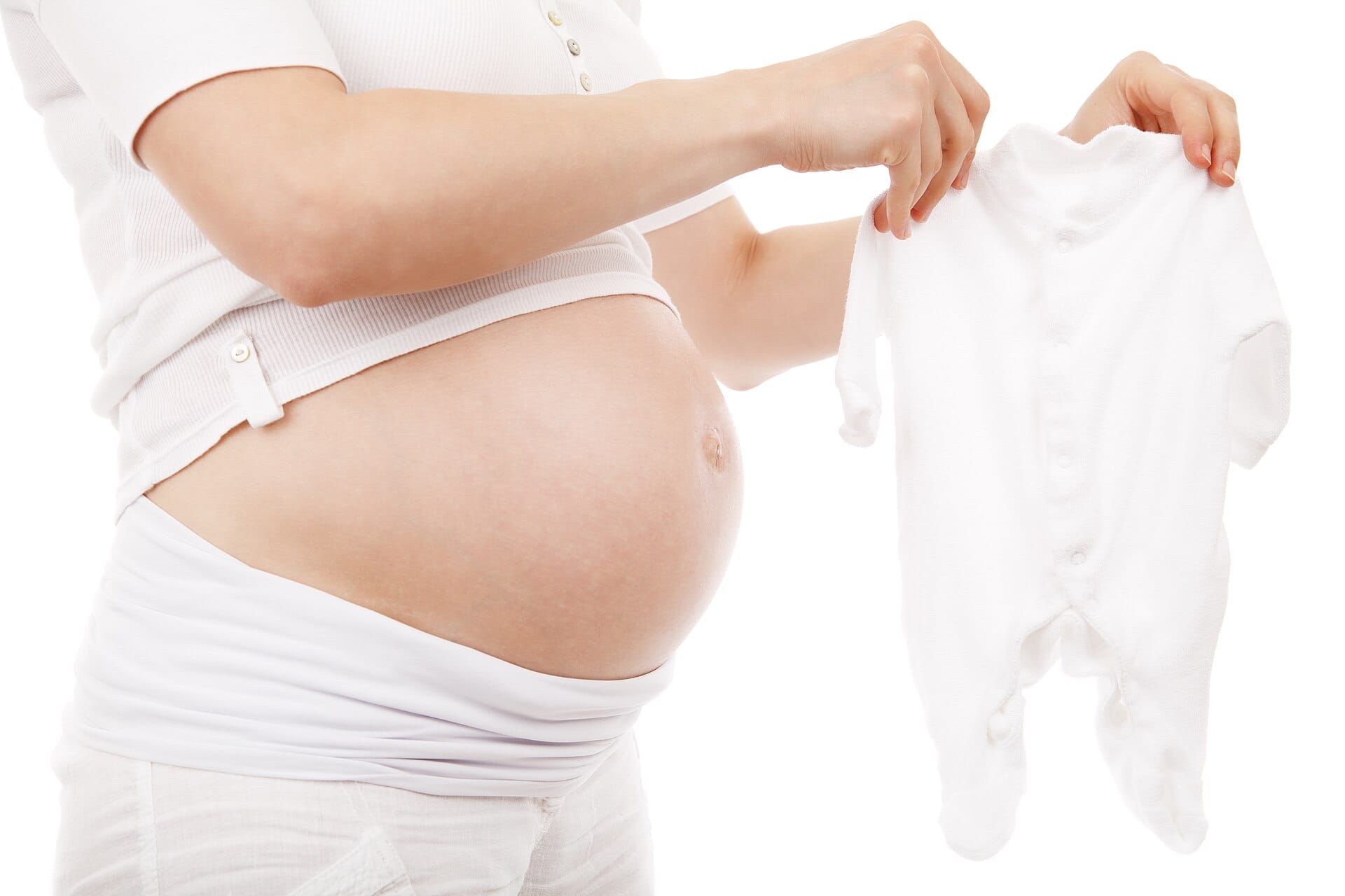 Zwangere vrouw in een wit shirt houdt een wit babyrompertje omhoog naast haar buik